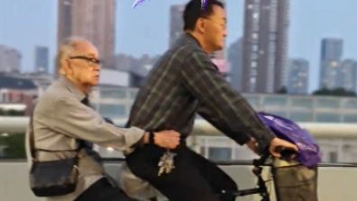 湖北武汉，街头一位中年男子骑着老式自行车，后座载着一位白发老人缓缓前行。